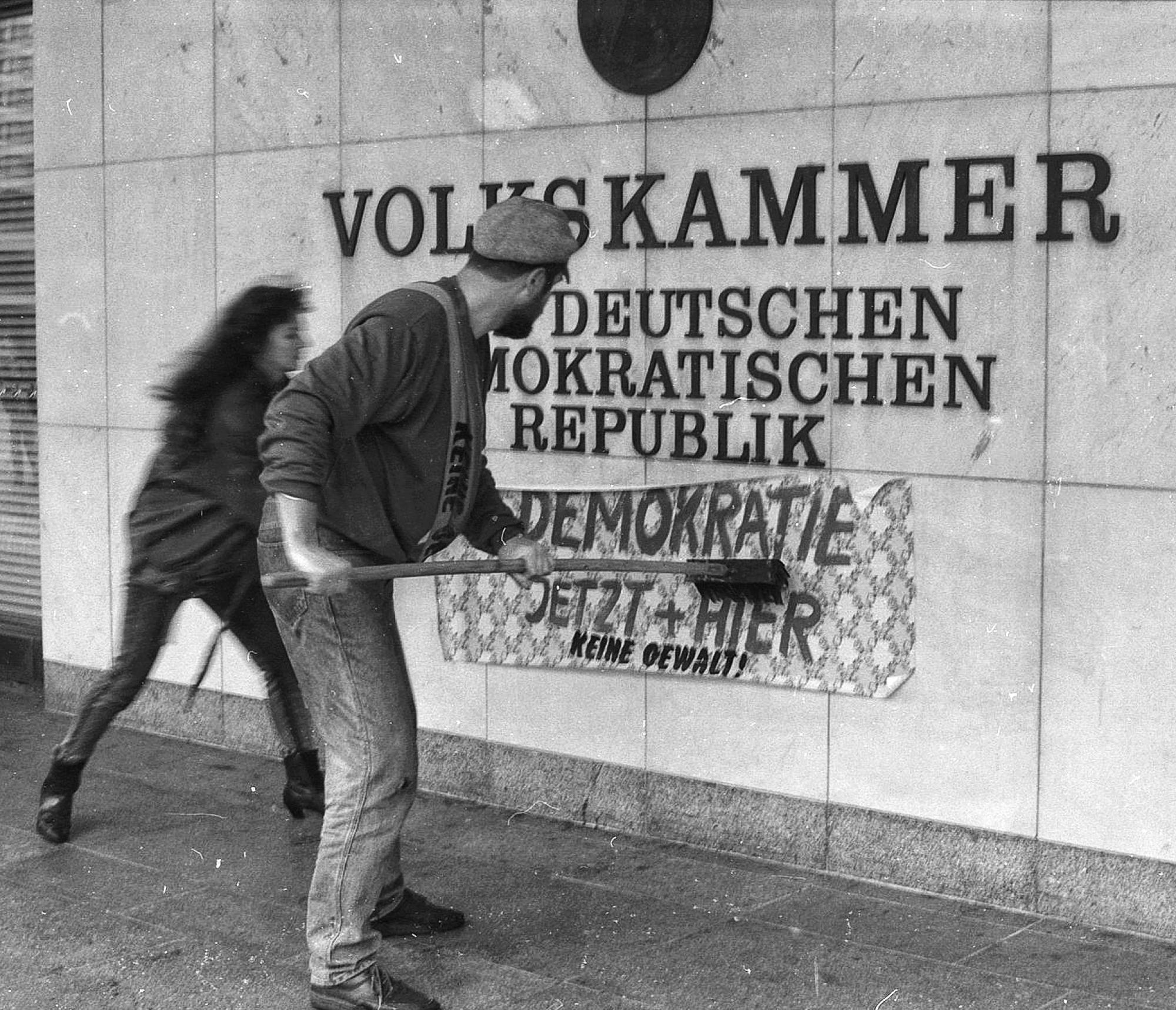 Zwei Demonstranten streichen eine neue Tapete unter das Schild der Volkskammer der DDR während der Alexanderplatz-Demonstration am 4. November 1989 in Ost-Berlin. Auf der Tapete steht "Demokratie jetzt und hier".