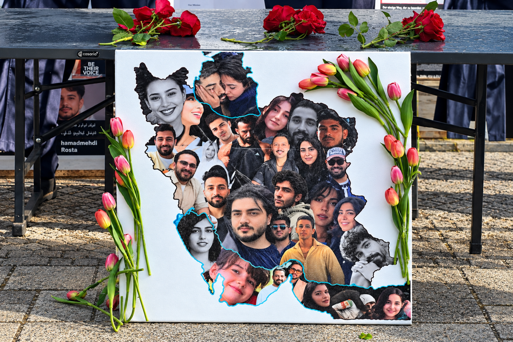 Ein Foto eines Protestschildes mit den Fotos von iranischen Menschen und Blumen darauf.
