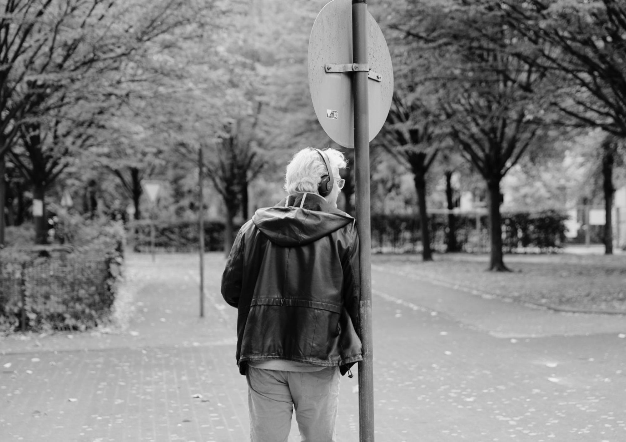 Eine Person mit Kopfhörern lehnt an einem Straßenschild. Sie ist von hinten fotografiert.